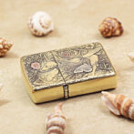 Brass Zippo-style lighter with intricate floral and moth engravings resting on sand among seashells.