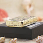 Gold Zippo lighter with engraved pattern resting on a black box, with seashells in the foreground.