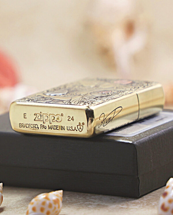 KZ452-05 Gold Zippo lighter with engraved pattern resting on a black box, with seashells in the foreground.
