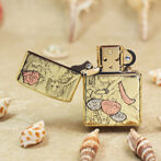 Gold-tone Zippo lighter with a floral and fruit engraving, open lid, resting on sandy beach among seashells.