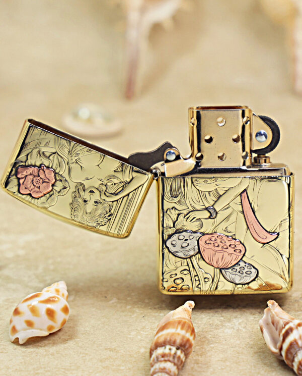 KZ456-02 Gold-tone Zippo lighter with a floral and fruit engraving, open lid, resting on sandy beach among seashells.
