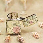 Gold decorative Zippo-style lighter with pink flower design, open lid, resting on sand with seashells in the foreground.