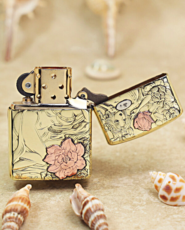 KZ456-03 Gold decorative Zippo-style lighter with pink flower design, open lid, resting on sand with seashells in the foreground.