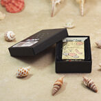 Zippo lighter with artwork in an Armor Case inside a black display box on a shell-strewn beach background.