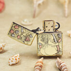 Gold-tone Zippo-style lighter with intricate engraved artwork, open lid, resting on a sandy beach with seashells nearby