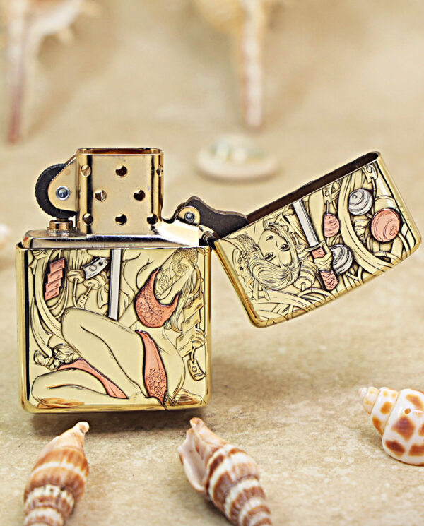KZ457-03 Gold-tone Zippo-style lighter with a pin-up girl engraving, open lid, resting on sand among seashells.
