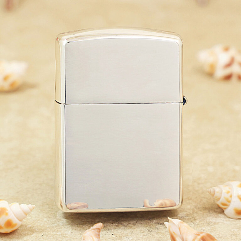 Shiny metal Zippo-style lighter standing upright on sand, surrounded by seashells nearby.