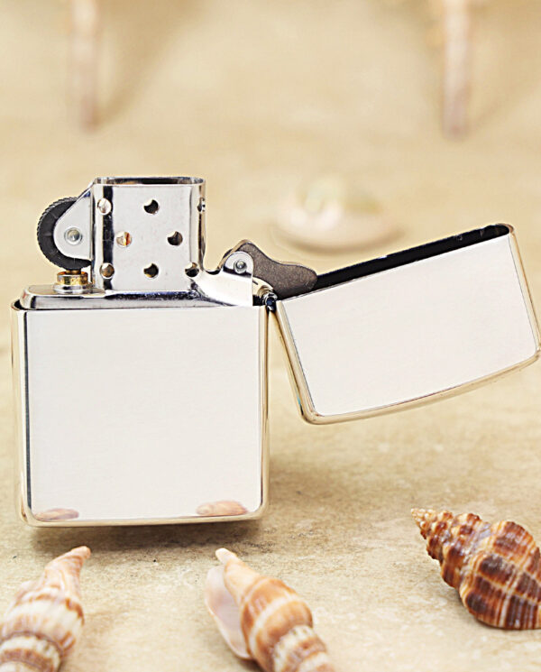 KZ461-03 Chrome Zippo-style lighter with its lid open, resting on sandy beach with scattered seashells nearby