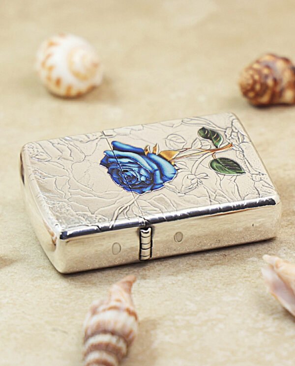 KZ461-04 Silver compact case with a blue rose engraving, placed on sand with seashells around it.