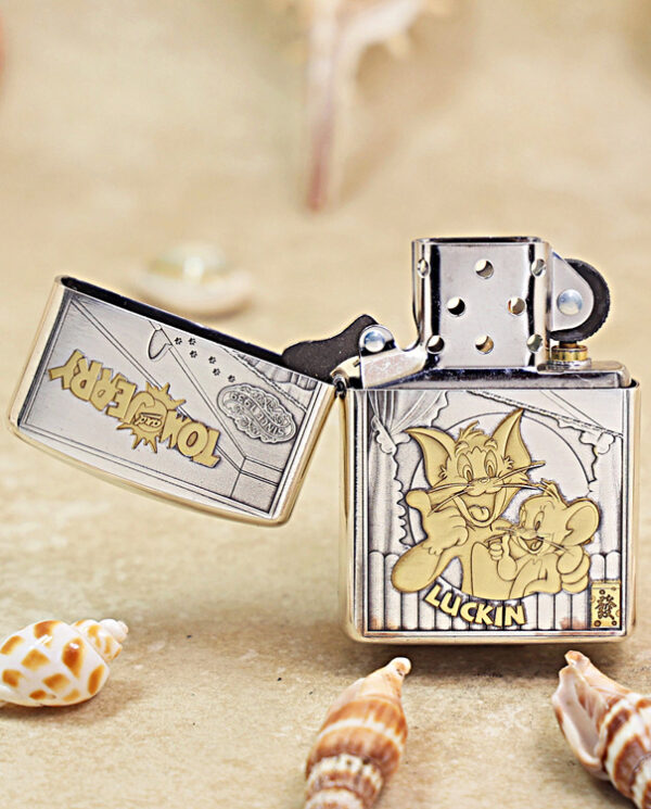 Zippo-style metal lighter open to show a Tom and Jerry illustration on the front cover, set on a sandy surface with seashells nearby