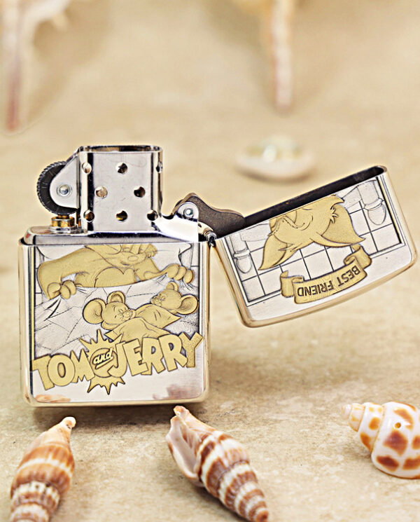 Open Tom and Jerry-themed Zippo lighter, metal case with cartoon panels resting on a sandy surface among seashells.