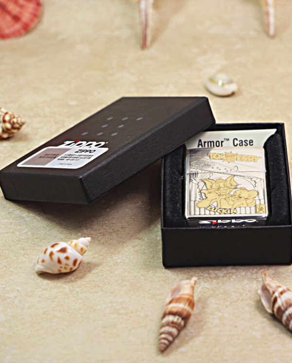 Open black presentation box showing a metal Tom and Jerry Armor Case with cartoon artwork inside on a sandy surface, surrounded by seashells.