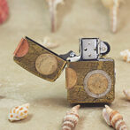 Zippo-style lighter with a vintage map design and attached coins, open lid, on a sandy beach with seashells nearby