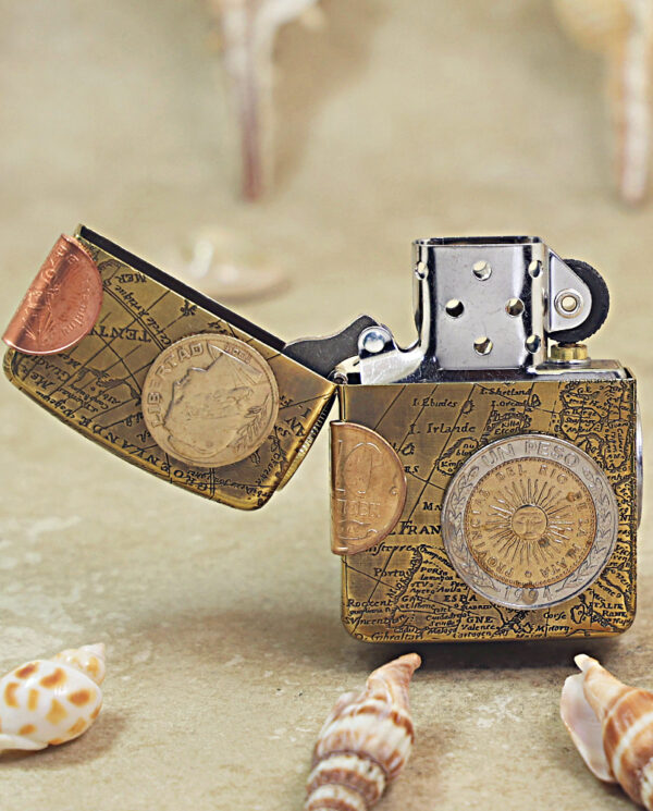 KZ468-02 Zippo-style lighter with a vintage map design and attached coins, open lid, on a sandy beach with seashells nearby