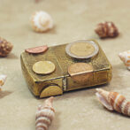 Small decorative gold box with coins arranged on its lid, surrounded by seashells on a sandy surface.