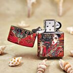 Decorative metal lighter with red Asian-inspired art showing a woman and a demon mask, lid open, on a sandy surface with shells nearby (informational product image).