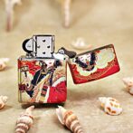 Metal lighter with a red and gold decorative Asian-inspired design, open lid, resting on a sandy surface with seashells nearby