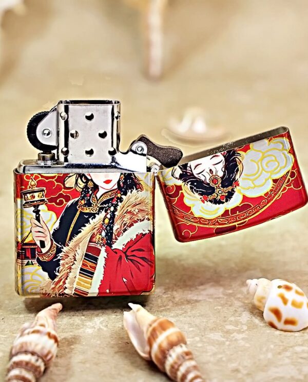 Metal lighter with a red and gold decorative Asian-inspired design, open lid, resting on a sandy surface with seashells nearby