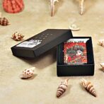 Small red decorative card inside an open black gift box, surrounded by seashells on a beige surface.