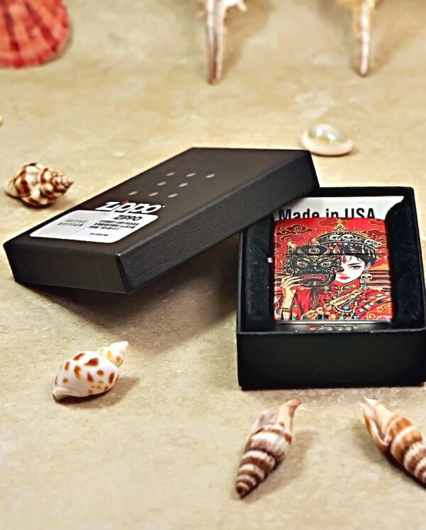 Small red decorative card inside an open black gift box, surrounded by seashells on a beige surface.