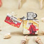 Colorful decorative lighter on sand, showing a red-cloaked traveler against a stylized landscape with seashells nearby.