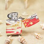 Colorful Zippo-style lighter with red and gold oceanic artwork, open lid, on a sandy beach with seashells nearby.