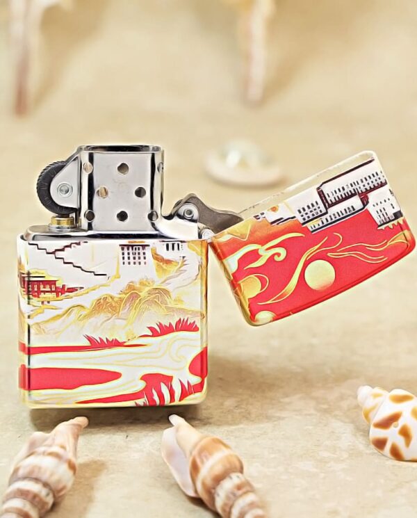 Colorful Zippo-style lighter with red and gold oceanic artwork, open lid, on a sandy beach with seashells nearby.