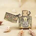 Gold engraved Zippo lighter with an Asian-inspired mask design, lid open, on a sandy surface with seashells nearby.