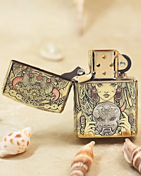 KZ501-02 Gold engraved Zippo lighter with an Asian-inspired mask design, lid open, on a sandy surface with seashells nearby.