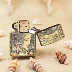 Gold engraved Zippo-style lighter with an ornate design and a woman's profile, lid open, on a sandy surface with seashells nearby.