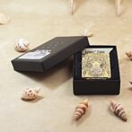 Open black box reveals a decorative gold card with intricate design, labeled Armor Case, on a sandy surface with seashells nearby.