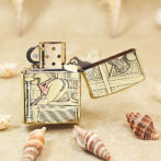 Brass flip-top lighter with ornate engraved panels resting on sand among seashells.