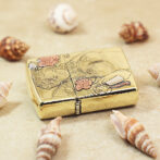 Gold decorative case with an engraved woman and floral design, placed among seashells on sand.