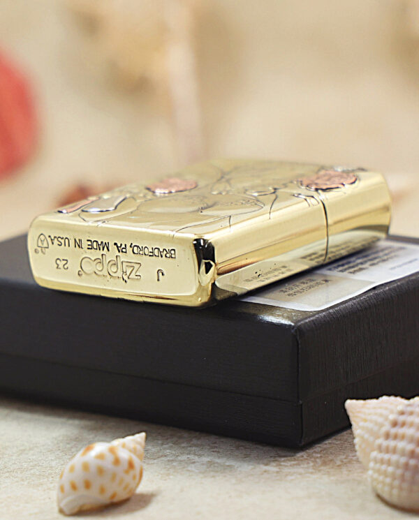 KZ506-05 Gold-tone Zippo lighter with a shell pattern rests on a black box, surrounded by seashells on a light surface.