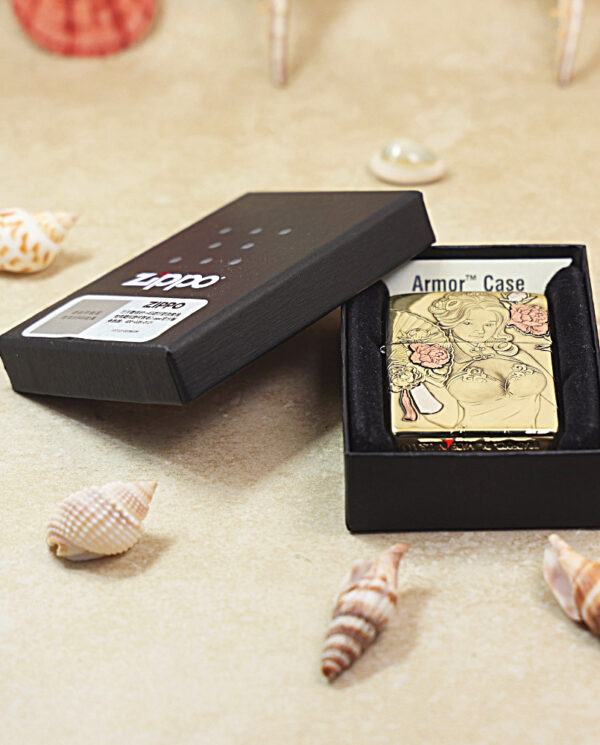 KZ506-07 Open black Zippo lighter box reveals a gold engraved lighter with floral and figure design, resting on a sandy beach with seashells nearby.
