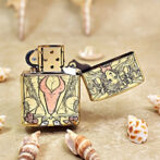 Gold-tone Zippo lighter with ornate tarot-style illustration of a stylized seated woman in a pink dress, open lid on a sandy surface with seashells nearby.