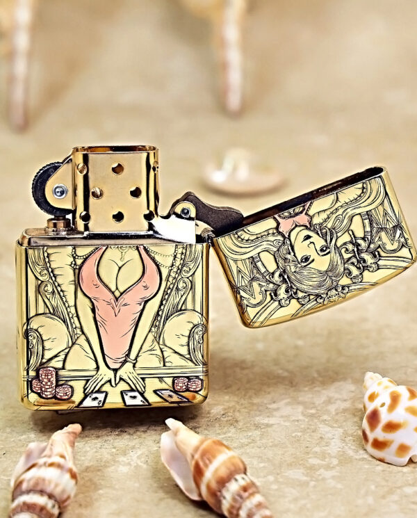 Gold-tone Zippo lighter with ornate tarot-style illustration of a stylized seated woman in a pink dress, open lid on a sandy surface with seashells nearby.