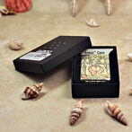 Zippo Armor Case lighter with illustrated woman, in a black box on a sandy surface with seashells scattered around.