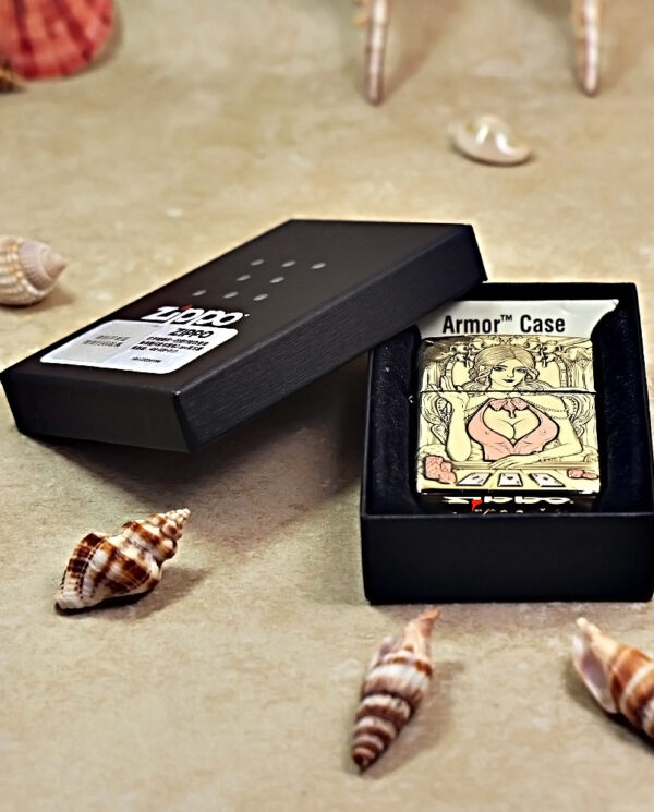 Zippo Armor Case lighter with illustrated woman, in a black box on a sandy surface with seashells scattered around.
