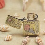 Decorative brass lighter with floral engraving, open lid, on a sandy surface with seashells nearby.