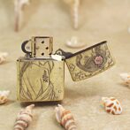 Vintage brass lighter with floral engravings, open lid, resting on sandy surface among seashells for decorative effect