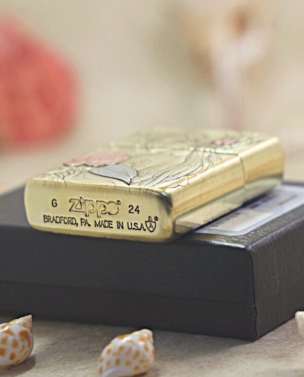 KZ509-05 Gold Zippo lighter with floral engravings resting on a black box, visible branding and 'Bradford, PA Made in USA'; small seashells scattered nearby.