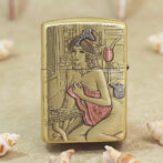 Gold-toned lighter engraved with a seated woman in a pink dress at a vanity against a geometric background.