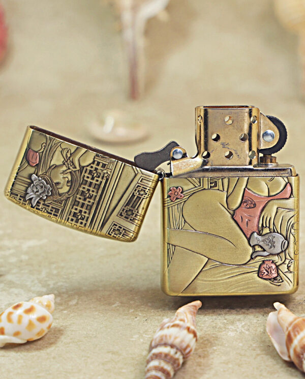 KZ510-02 Gold-tone Zippo lighter with intricate art-deco panels, open lid, resting on a sandy surface among seashells.