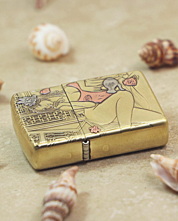 KZ510-04 Gold-tone lighter with intricate engravings of a reclining figure and floral patterns, placed on a sandy surface among seashells.