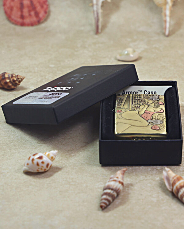 KZ510-06 Open black box reveals a gold Zippo lighter with a decorative design, placed on a sandy surface with seashells nearby.