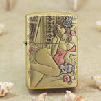 Gold-tone Zippo lighter with an engraved art-deco image of a lounging woman in a pink swimsuit and flower in her hair, set among decorative patterns.