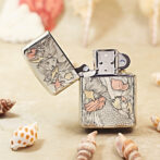 Metal Zippo-style lighter with a detailed mermaid and seashell engraving, open lid, on a sandy surface with shells nearby.