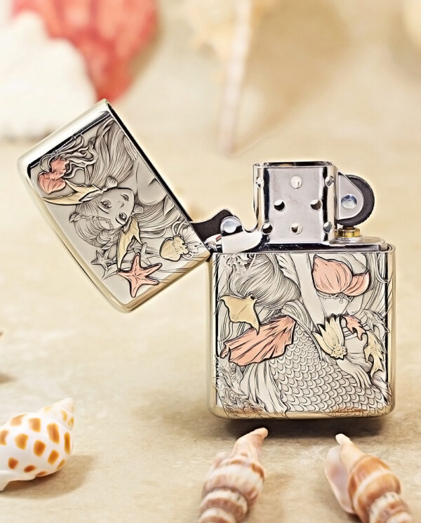 Metal Zippo-style lighter with a detailed mermaid and seashell engraving, open lid, on a sandy surface with shells nearby.