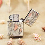 A metal Zippo lighter with an intricate koi fish engraving and an open lid, placed on a sandy surface with seashells nearby.